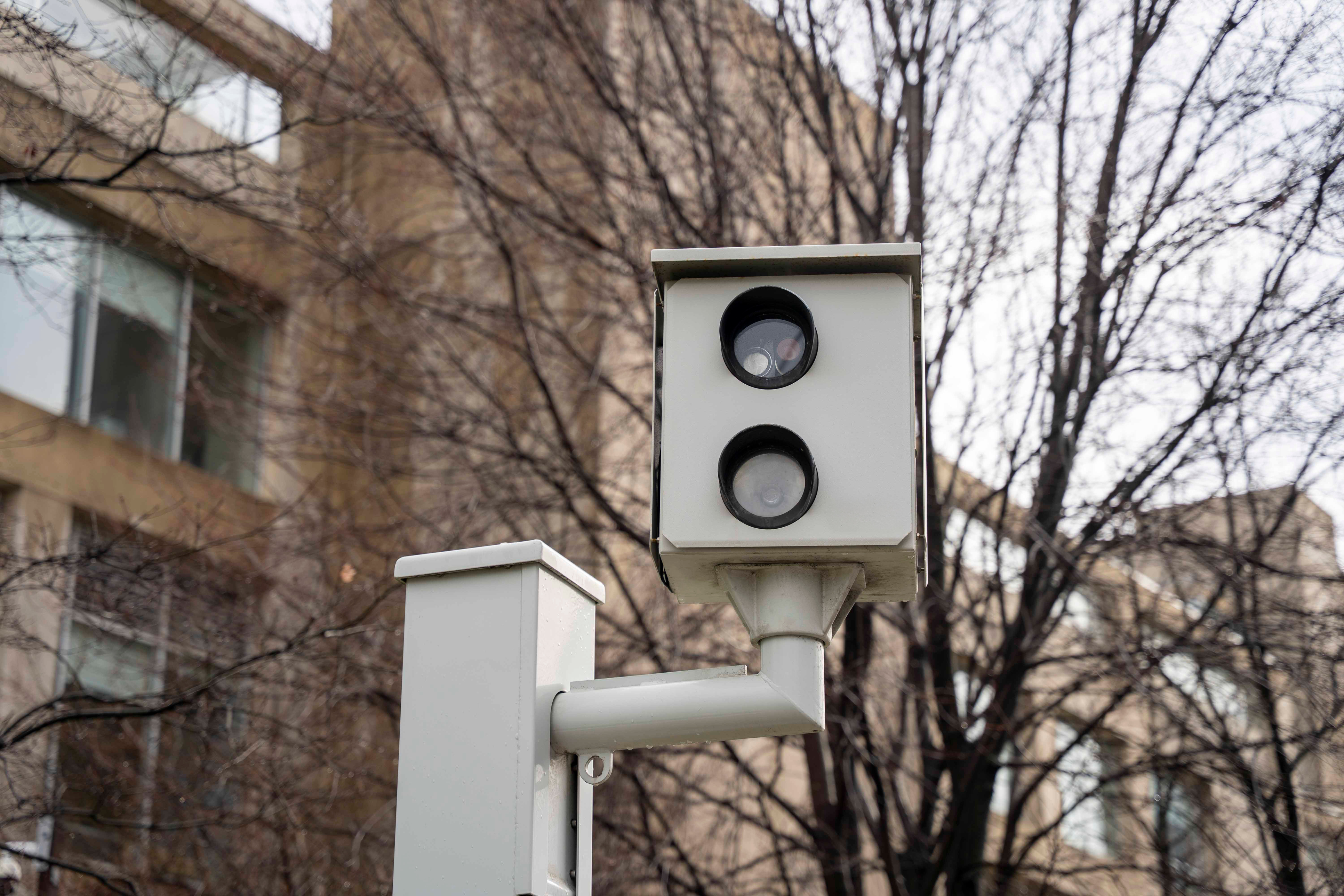 Close-up of a roadside speed camera mounted on a pole, used for automated traffic enforcement in a residential area of Toronto with buildings and bare trees in the background.